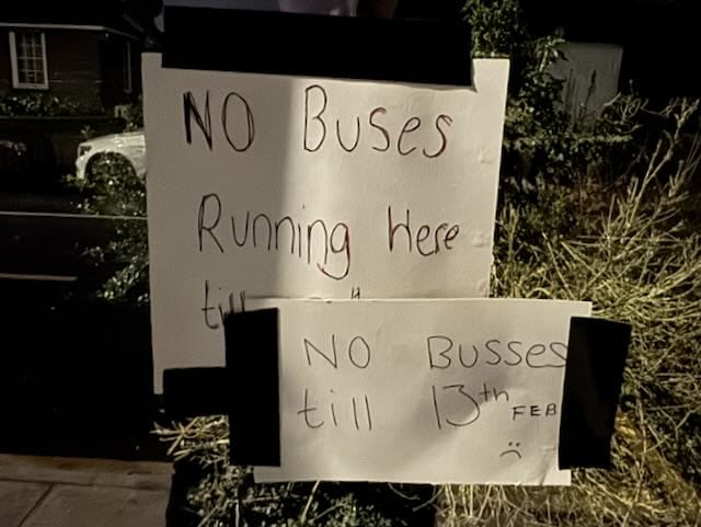 handwritten sign saying No Busses till 13th, gaffer-taped over an older sign saying No Buses Running Here, on a glass bus shelter
