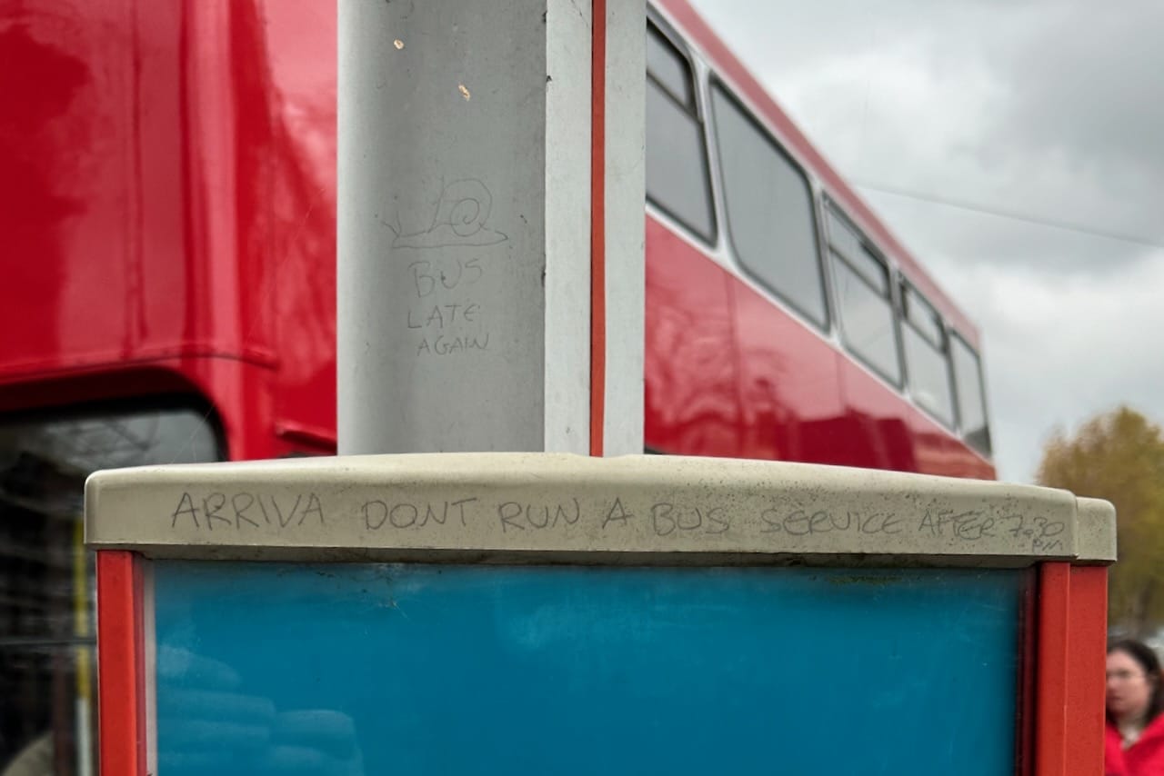 Foreground: graffiti on a bus stop pole: a drawing of a snail, bus late again, Arriva don't run a bus service after 7.30 pm. Background: a red MCW Metrobus